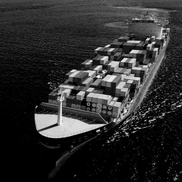 Zwart-witfoto van Luc Huysmans van een groot vrachtschip gevuld met talloze zeecontainers, dat door kalm water beweegt en een schaduw werpt op het zeeoppervlak.