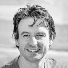 Rafaël Mondre, een glimlachende blanke man met kort, warrig haar, poseert in dit zwart-witportret. Als hij buiten staat met een onscherpe natuurlijke achtergrond, draagt hij een lichte, casual outfit