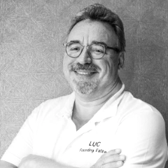Een zwart-wit portret van een glimlachende man van middelbare leeftijd met bril en sikje, gekleed in een poloshirt met de tekst 'Rafaël Mondre' en 'grondlegger' in reliëf. De achtergrond