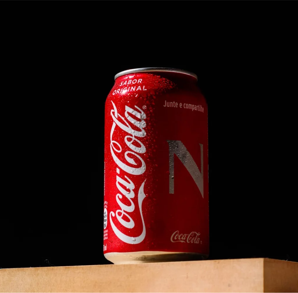 Een coca-colablikje staat op een houten oppervlak tegen een donkere achtergrond en is aan één kant verlicht, wat de felrode kleur en het opvallende witte logo benadrukt. een gedeeltelijke schaduw werpt over het blik, waardoor de textuur ervan wordt versterkt.