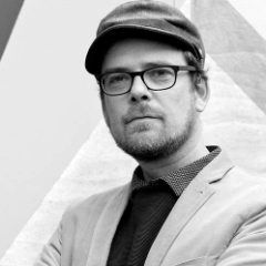 Zwart-wit portret van Rafaël Mondre, een man met een bril, een platte pet en een pak met overhemd en stropdas. Hij heeft een korte baard en kijkt recht in de ogen