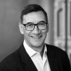 Een zwart-wit portret van een glimlachende man van middelbare leeftijd met een bril, een donkere blazer en een wit overhemd. Hij heeft kort grijzend haar en staat voor een wazige architecturale achtergrond