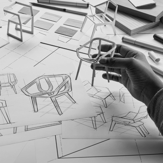 Iemands hand houdt een schaalmodel vast van een moderne stoel ontworpen door Michiel Goethals boven een bureau vol bouwtekeningen en schetsen van verschillende stoelontwerpen, naast boeken en tekengereedschappen.
