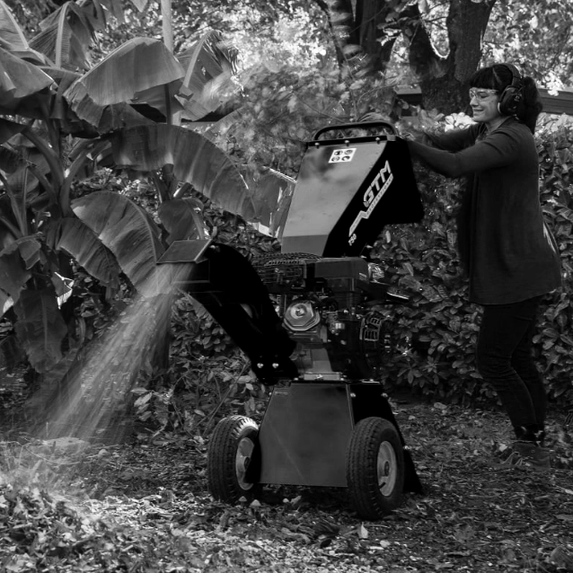 Christina De Wild bedient met een koptelefoon een grote houtversnipperaar in een weelderige tuin en steekt takken in die versnipperd moeten worden. De machine werpt mulch uit, omringd door dicht gebladerte terwijl zonlicht erdoorheen filtert