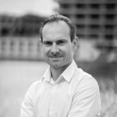 Een zwart-wit portret van Glenn Van Croonenborch glimlachend, met kort haar, gekleed in een wit overhemd met knoopjes, staande buiten met vage gebouwen en bomen op de achtergrond.