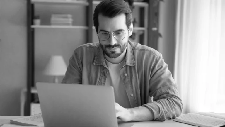 Een zwart-wit beeld van een gefocuste jongeman met bril en baard, gekleed in een casual shirt, werkend aan een nieuwe website aan een bureau omringd door boeken in een goed verlichte kamer.