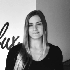 Een professionele portretfoto van Romy Lupo, een jonge vrouw met lang, steil haar en een zwart topje. Ze staat tegen een neutrale achtergrond en kijkt met een subtiele glimlach rechtstreeks in de camera