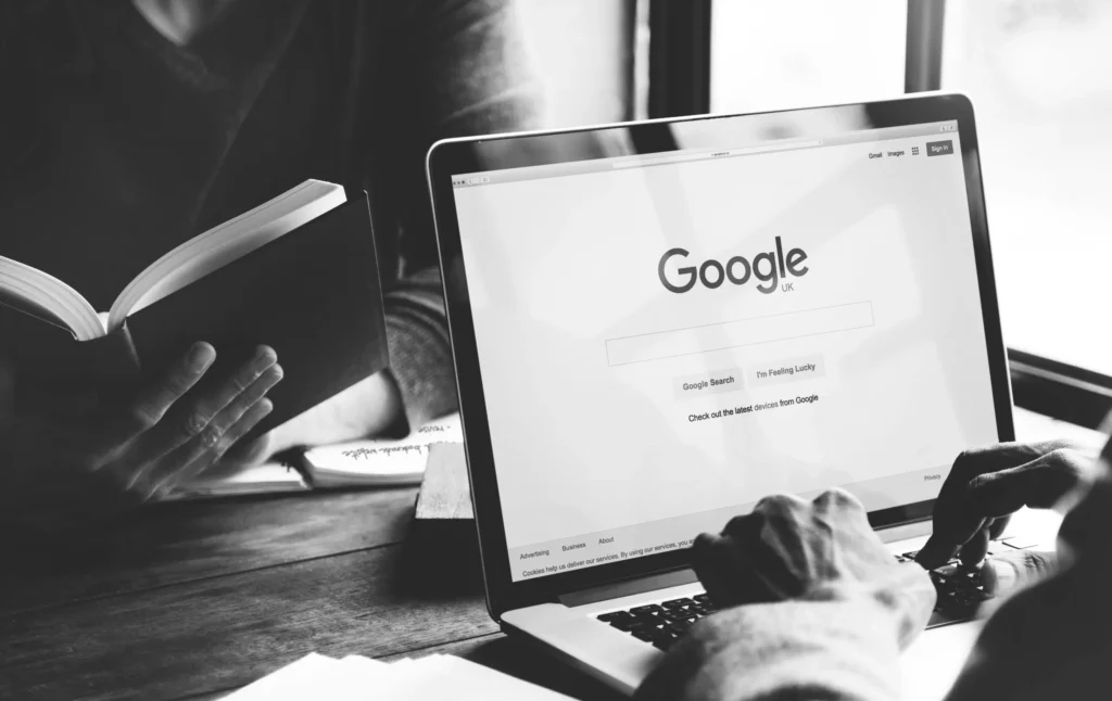 Een zwart-wit beeld van twee individuen aan een houten tafel; de één leest een boek, terwijl de ander een laptop gebruikt waarop de startpagina van Google Zoeken wordt weergegeven.