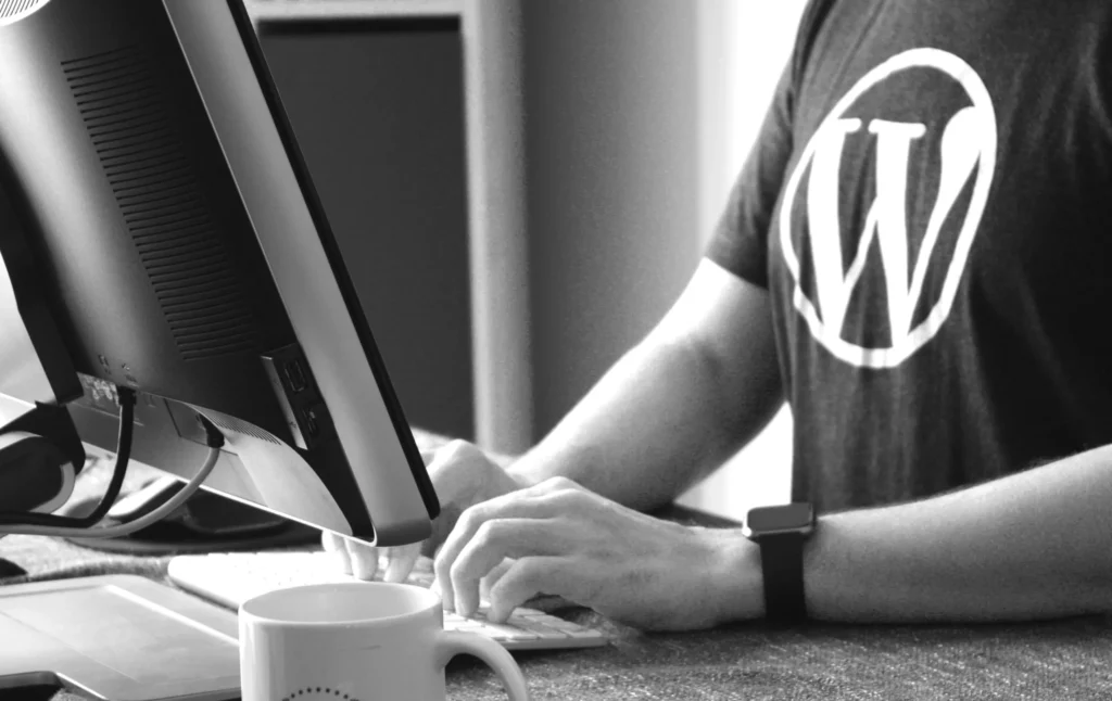 Een persoon die een grijs T-shirt met een WordPress-logo draagt, werkt op een computer en typt op een toetsenbord met de linkerhand zichtbaar. een witte mok en een smartphone rusten op het bureau naast de monitor. de afbeelding is in zwart-wit.