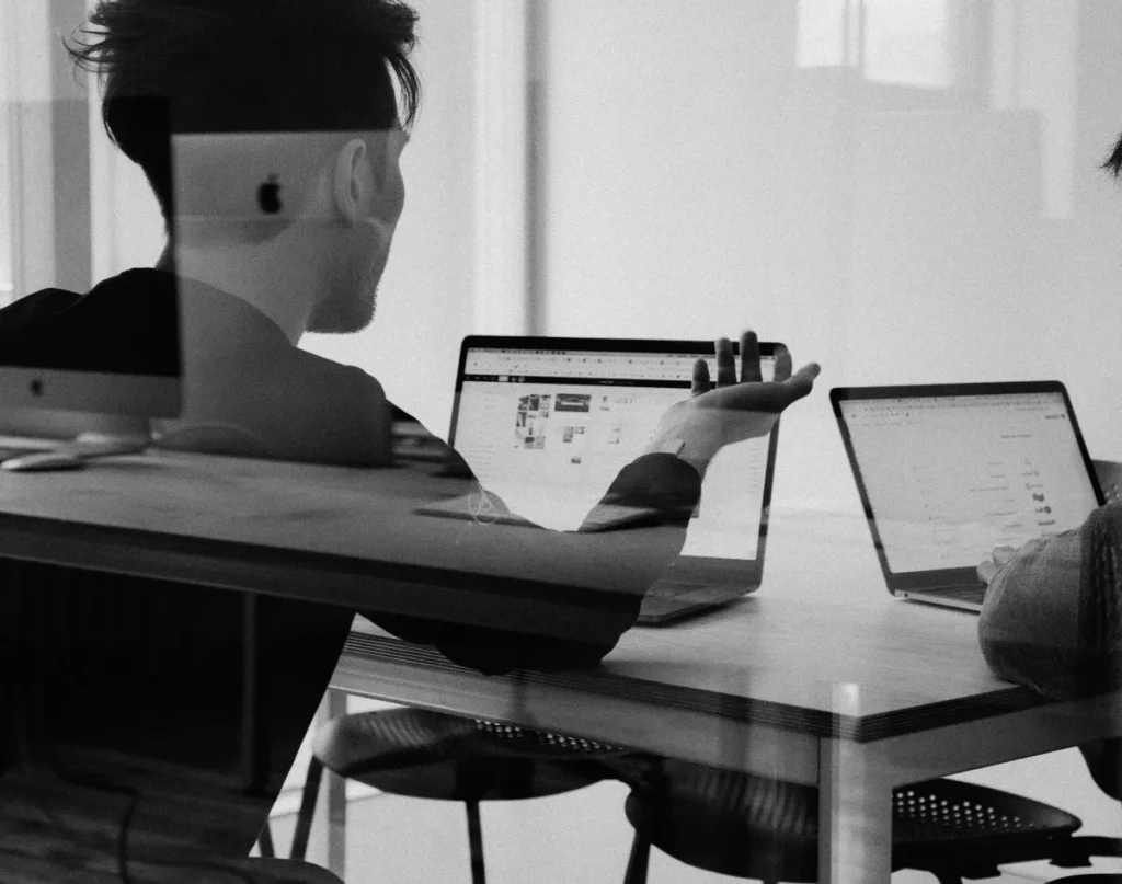 Een zwart-witfoto van een jonge man met een bril, zittend aan een bureau met twee laptops open voor hem. hij gebaart met één hand, schijnbaar in gesprek met iemand aan de andere kant van de tafel.