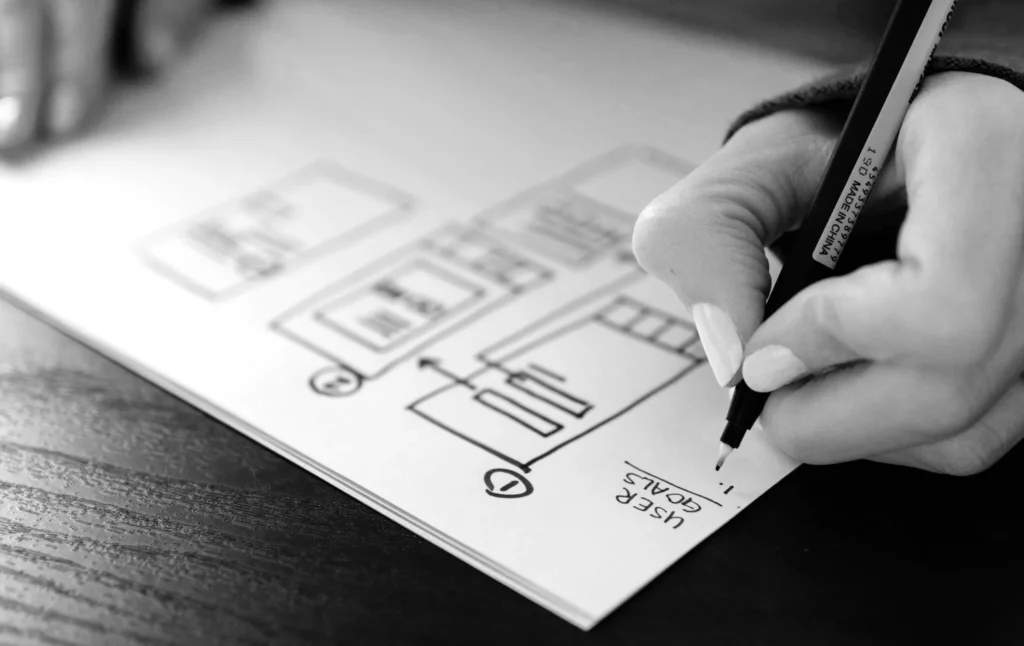 Een zwart-witafbeelding van de hand van een persoon die een pen vasthoudt en een draadframe schetst voor een website-ontwerp op papier, waarbij de nadruk ligt op lay-outelementen zoals kopteksten en pictogrammen.