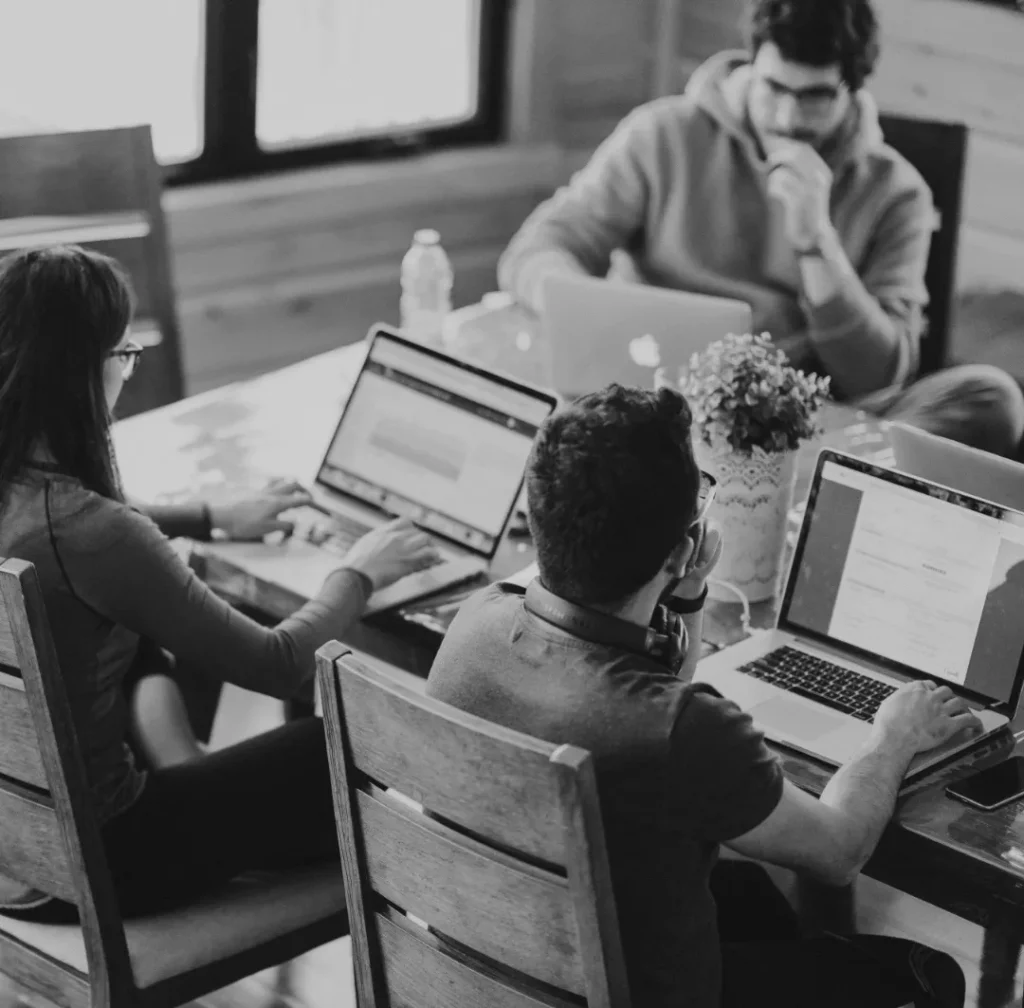 Vier mensen werken samen in een kantooromgeving, met behulp van laptops rond een houten tafel. ze zijn bezig met werk met gerichte uitdrukkingen. de omgeving is informeel en goed verlicht, met een moderne inrichting.