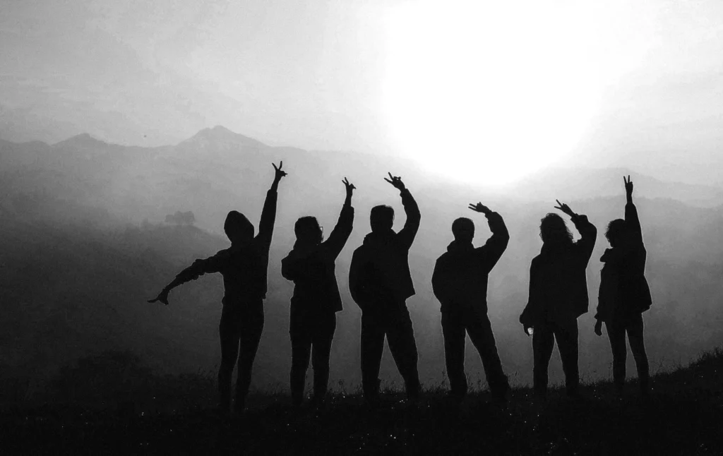 Silhouetten van zes mensen die op een heuvel staan, met opgeheven armen in verschillende vrolijke gebaren, tegen een heldere, gloeiende zon met zachte bergcontouren op de achtergrond.