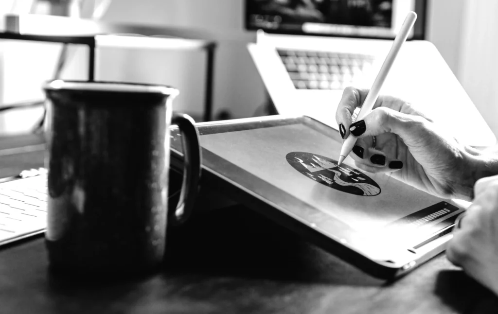 Een zwart-witafbeelding van de hand van een persoon die een stylus vasthoudt boven het scherm van een digitale tablet met een afbeelding, met een laptop en een koffiemok vlakbij op een bureau.