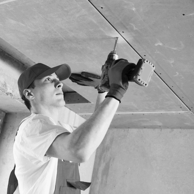 Een bouwvakker in een wit t-shirt en pet gebruikt een boormachine om een plafondpaneel vast te zetten. Hij is geconcentreerd op zijn taak gericht en werkt binnenshuis in een bouwomgeving. Het zwart en wit