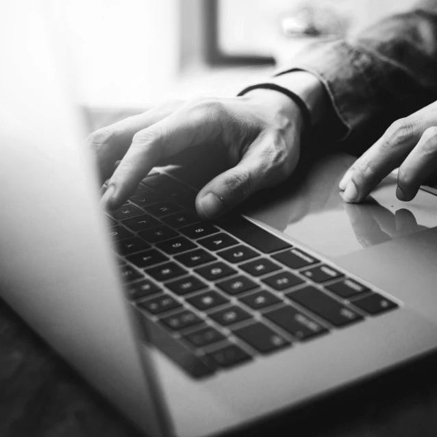 Close-up, zwart-witfoto van Glenn Van Croonenborch's handen die typen op het toetsenbord van een laptop, met een onscherpe achtergrond die het toetsenbord en de handen benadrukt.