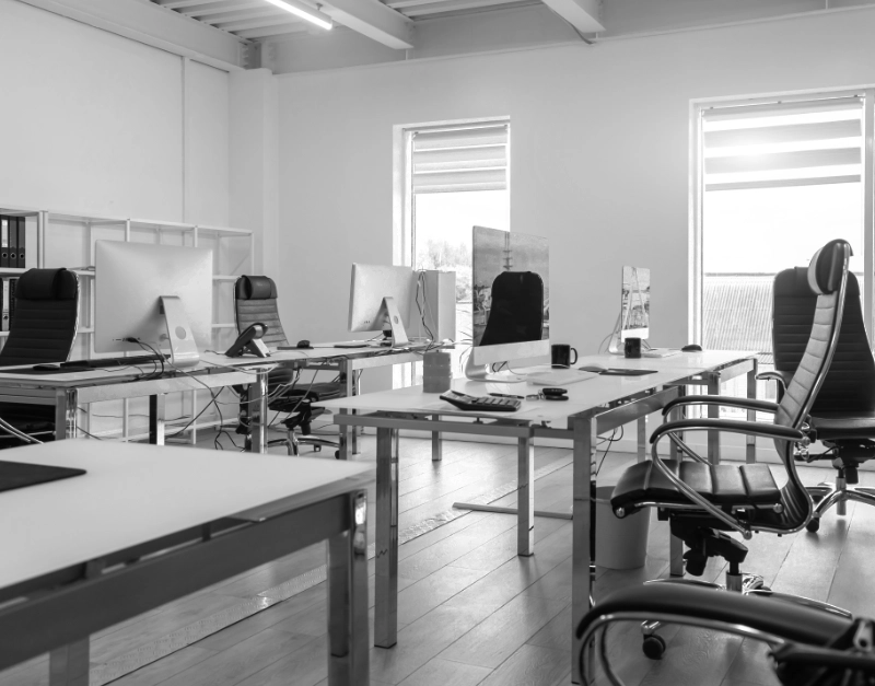 Een moderne kantoorruimte in zwart-wit met diverse bureaus met computers, ergonomische stoelen en twee grote ramen met gedeeltelijk getrokken zonwering. de kamer is goed verlicht en ziet er georganiseerd en leeg uit.