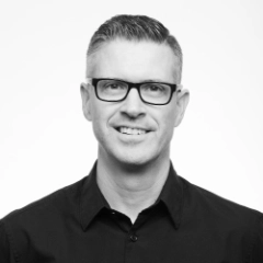 Zwart-wit portret van een glimlachende man met kort haar, een bril en een zwart shirt. Stefan Vermeulen heeft een strak uiterlijk en kijkt rechtstreeks in de camera.