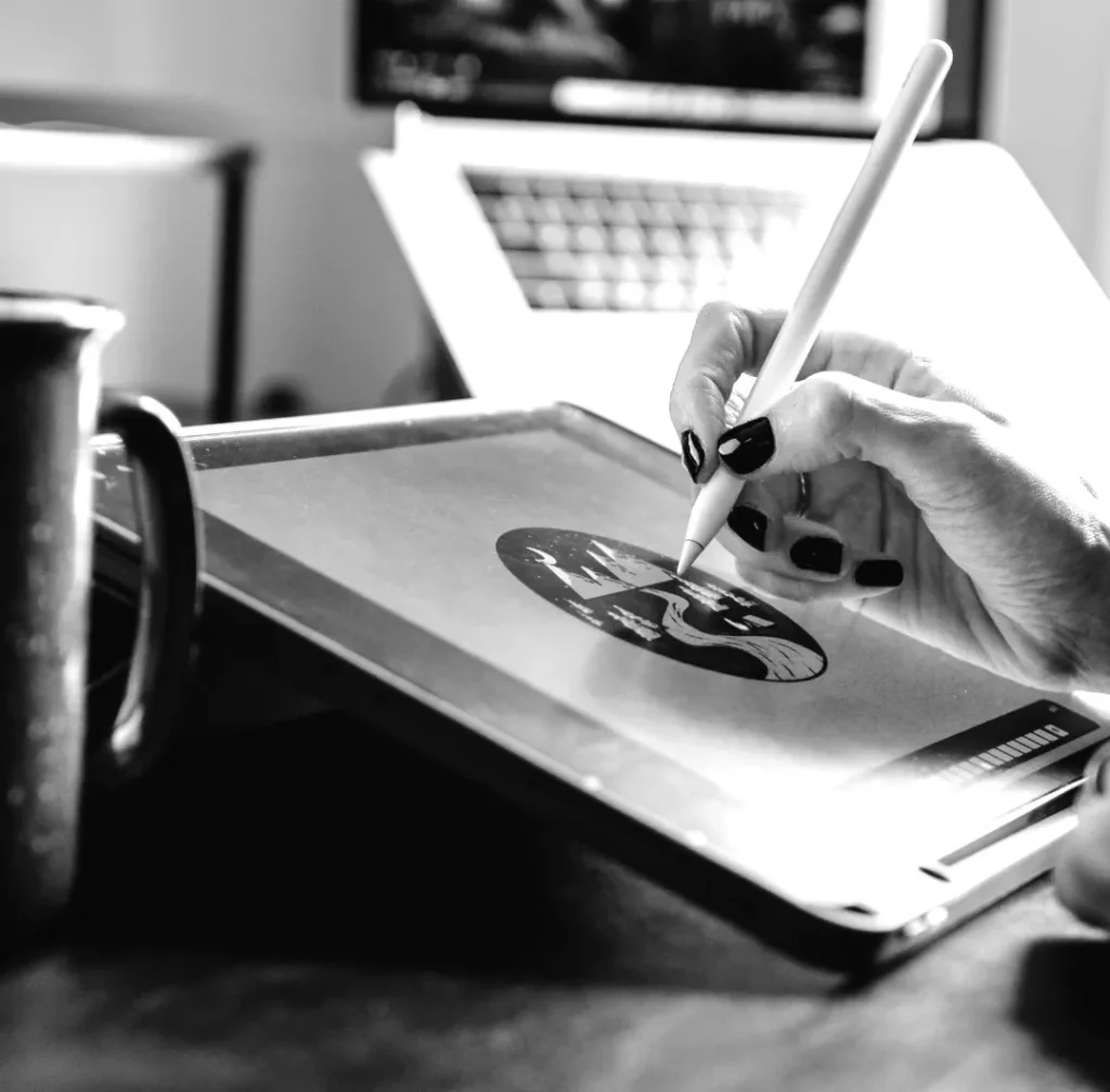 Een zwart-wit afbeelding van iemands hand die een stylus gebruikt om op een digitaal tablet te tekenen, met een laptop op de achtergrond op een bureau.