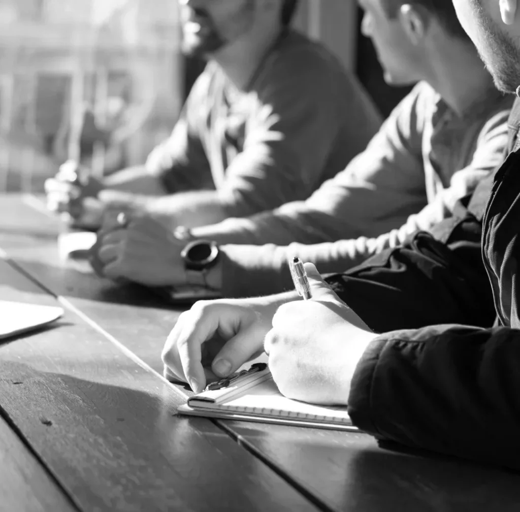 Drie personen concentreerden zich op het schrijven van aantekeningen aan een tafel tijdens een vergadering, geaccentueerd door zonlicht dat door een raam naar binnen scheen, waardoor een contrastrijk zwart-witeffect ontstond.