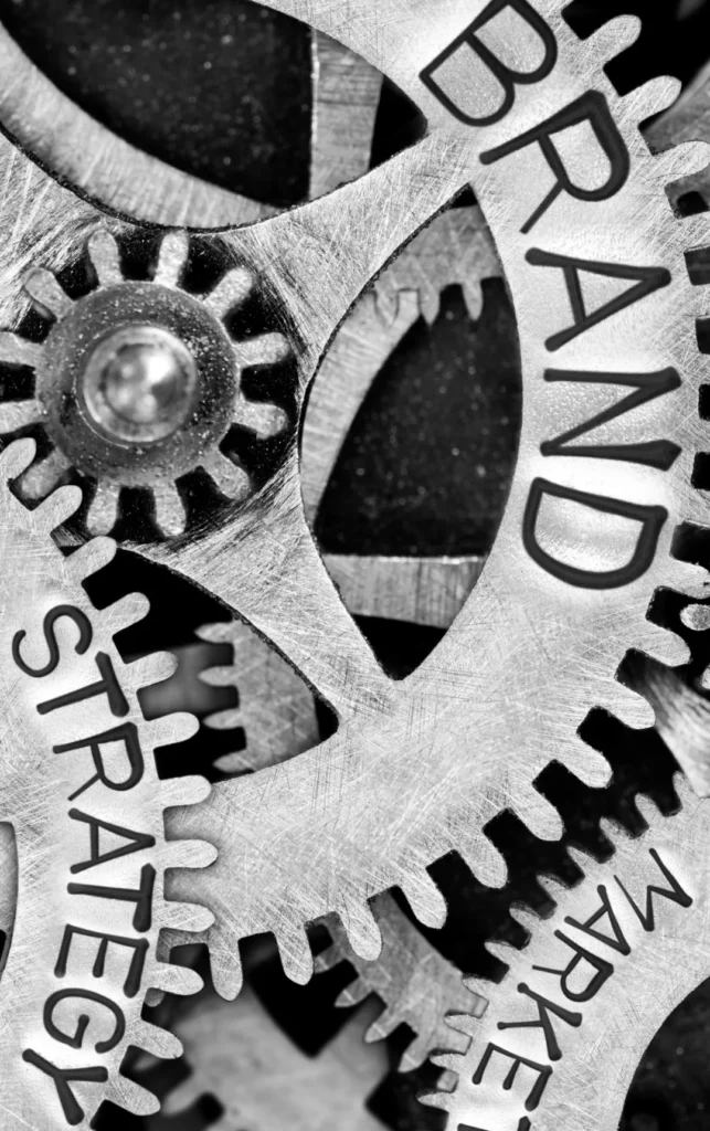 Close-up zwart-wit beeld van in elkaar grijpende metalen tandwielen met de woorden 'merk', 'strategie' en 'ervaring' erop gegraveerd, die de onderling verbonden elementen van de bedrijfsstrategie symboliseren.