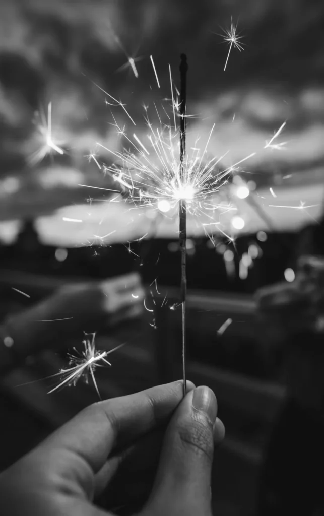 Een zwart-wit afbeelding van een hand die een verlicht sterretje vasthoudt. heldere vonken spatten uit het sterretje met subtiel wazig licht op de achtergrond, wat een gevoel van feest of feestelijkheid geeft.