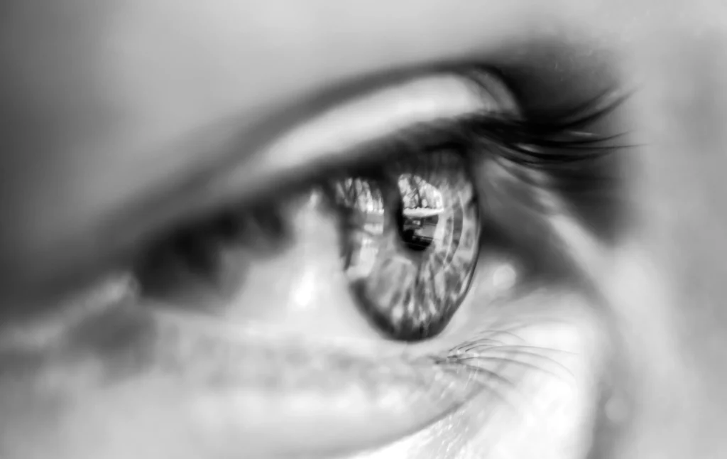 Close-up zwart-wit beeld van een menselijk oog, met de nadruk op de gedetailleerde iris en wimpers. reflecties die mogelijk gebouwen laten zien, zijn zichtbaar in de pupil, waardoor diepte en intriges aan de foto worden toegevoegd.