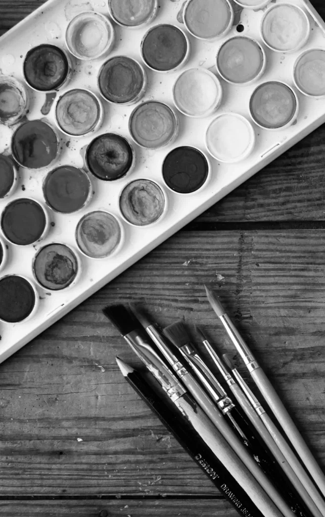 Een zwart-witfoto van een gebruikt aquarelverfpalet met meerdere kleuren naast een set penselen met verschillende punten, liggend op een houten oppervlak. de artistieke gereedschappen vertonen tekenen van veelvuldig gebruik.