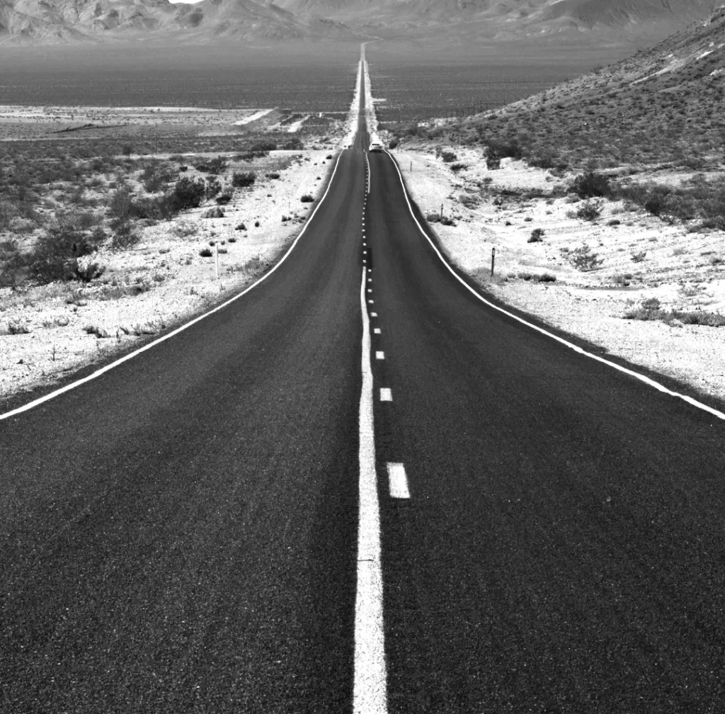 Zwart-wit beeld van een lange rechte weg die zich uitstrekt tot aan de horizon door een dor woestijnlandschap met aan weerszijden schaarse vegetatie. een heldere hemel en de weg gemarkeerd met een stippellijn zijn zichtbaar.