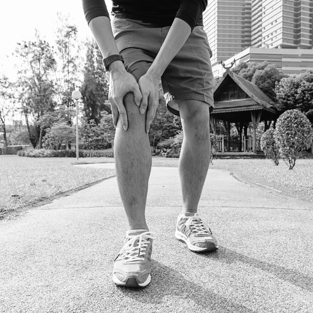 Een zwart-wit beeld van een persoon die zijn knie vasthoudt terwijl hij op een parkpad staat, waarbij alleen zijn onderlichaam, gekleed in korte broek en hardloopschoenen, te zien is, met gebouwen en groen op de achtergrond wazig
