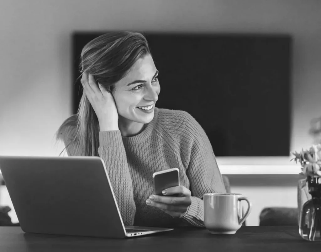 Een vrouw glimlacht terwijl ze naar het scherm van haar laptop kijkt, terwijl ze een smartphone in één hand houdt. ze zit aan een tafel met een mok en bloemen, in een warm verlichte kamer. de afbeelding is in zwart-wit.