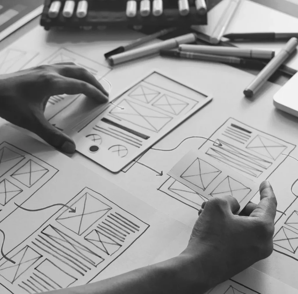 Een zwart-witafbeelding van de handen van een persoon die werkt aan draadschetsen verspreid over een tafel, met verschillende tekengereedschappen en een rekenmachine in de buurt.